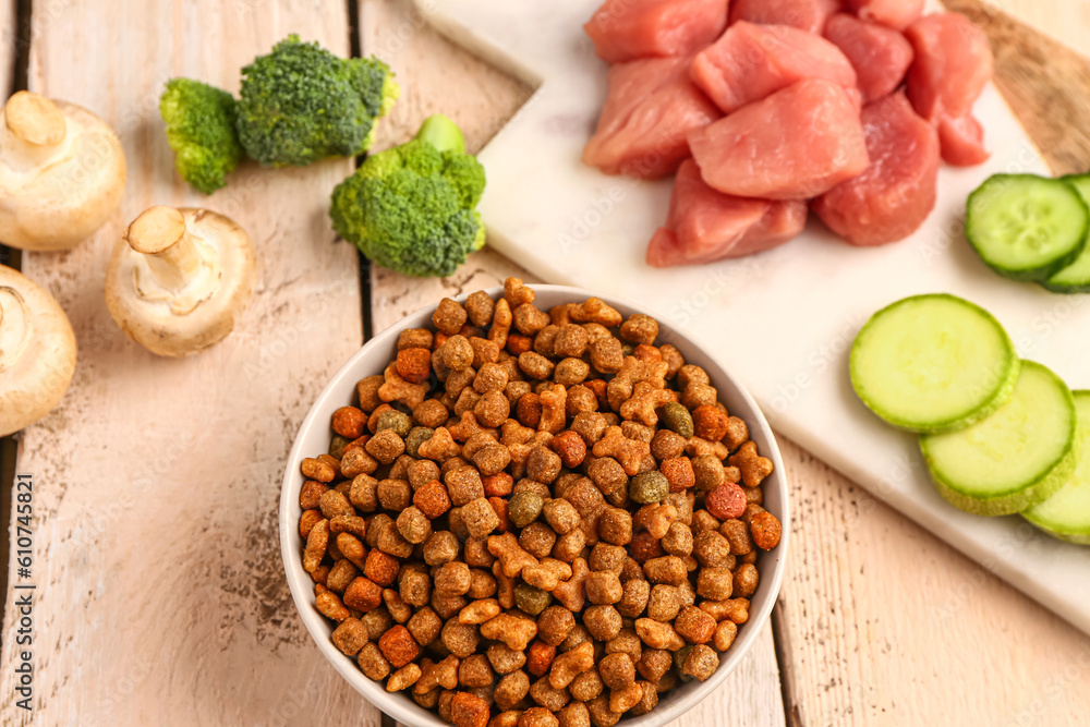 Bowl with dry pet food, raw meat and natural products on light wooden background, closeup