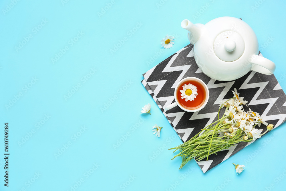 Teapot with cup of natural chamomile tea and flowers on blue background