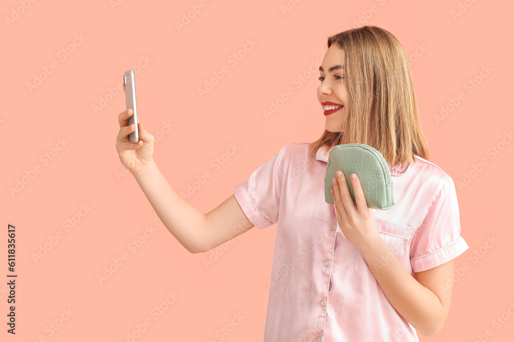 Young woman with cosmetic bag and mobile phone taking selfie on pink background