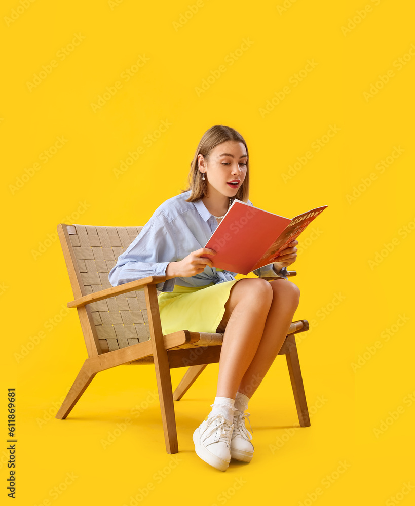 Young woman reading magazine in wooden armchair on yellow background