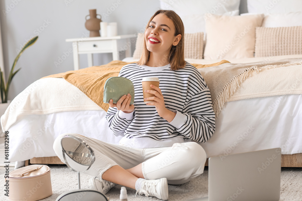 Young woman with cosmetic bag and cup of coffee sitting in bedroom