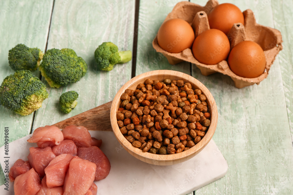 Bowl with dry pet food, raw meat, eggs and broccoli on color wooden background, closeup