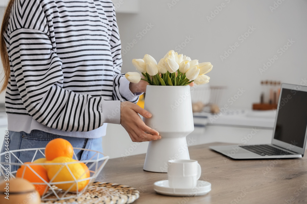 Woman putting bouquet of white tulip flowers into vase on wooden table in kitchen