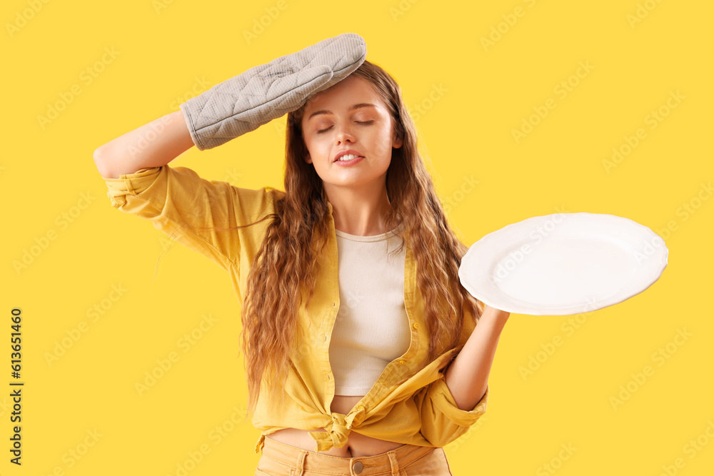 Tired young woman with empty plate and oven mitten on yellow background
