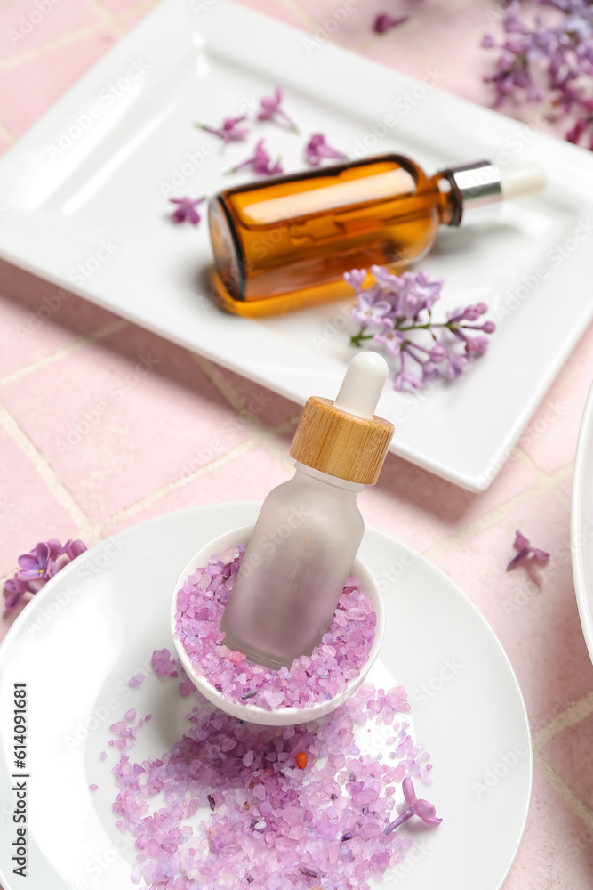 Bottles of lilac essential oil, sea salt and flowers on pink background, closeup