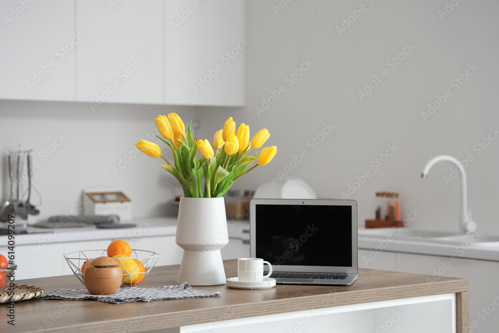 Vase with yellow tulip flowers, modern laptop, fruits and cup of coffee on wooden table in light kit