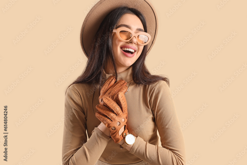 Stylish young woman in sunglasses and leather gloves on beige background