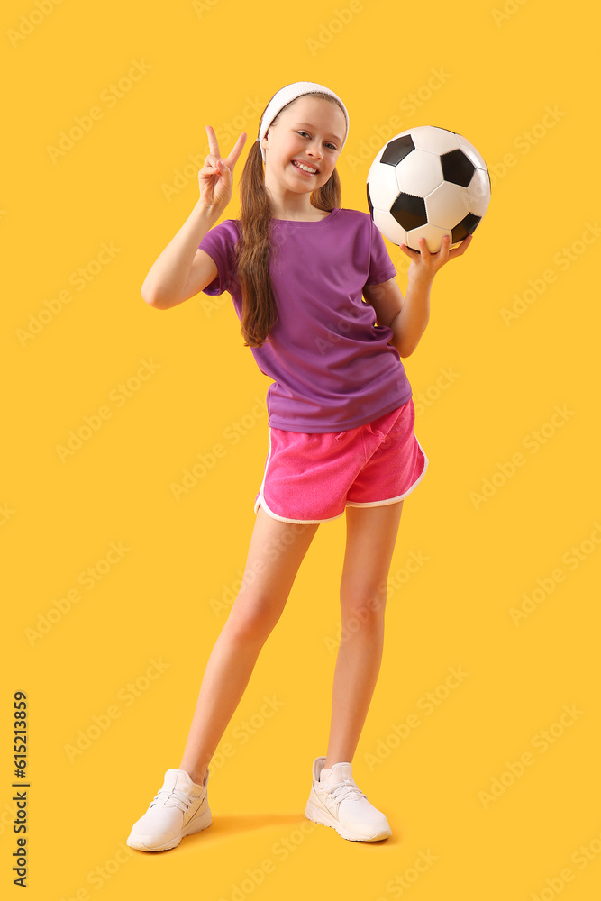 Sporty little girl with soccer ball showing victory gesture on yellow background