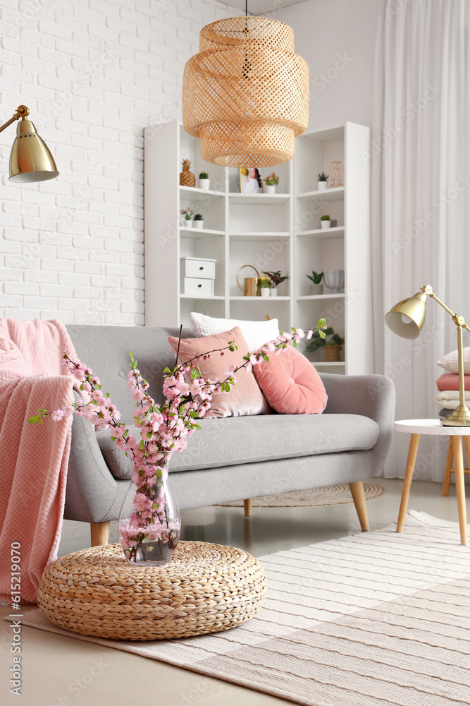 Vase with blooming sakura branches near grey sofa in interior of living room