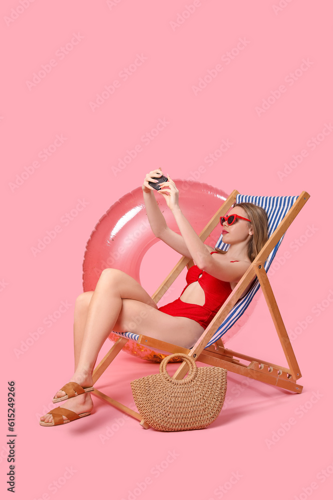 Young woman with beach accessories taking selfie in deck chair on pink background