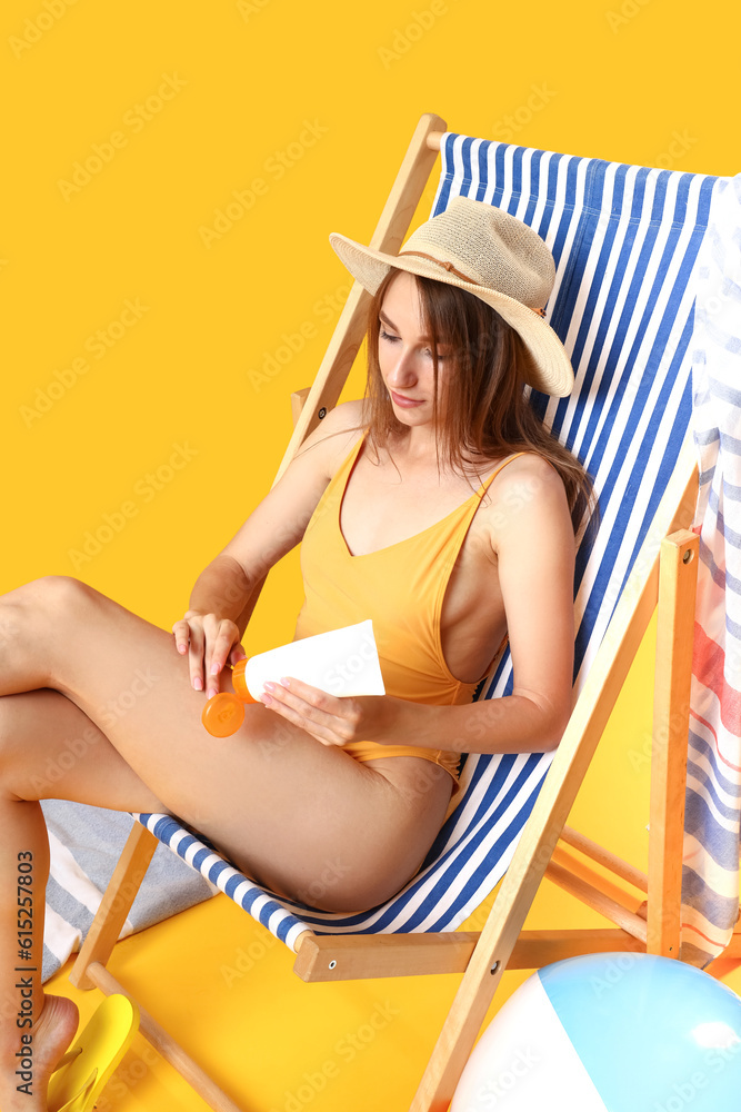Young woman applying sunscreen cream in deck chair on yellow background