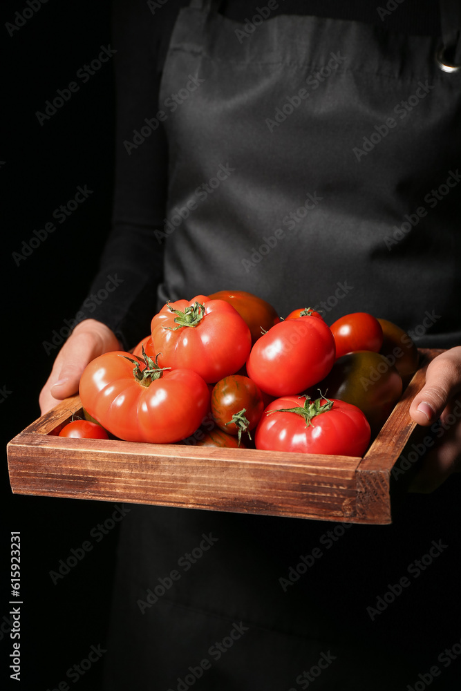 Woman holding wooden box with different fresh tomatoes on black background, closeup