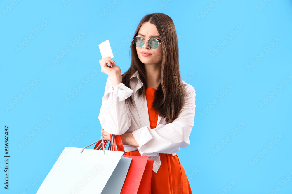 Thoughtful young woman with gift card and shopping bags on blue background