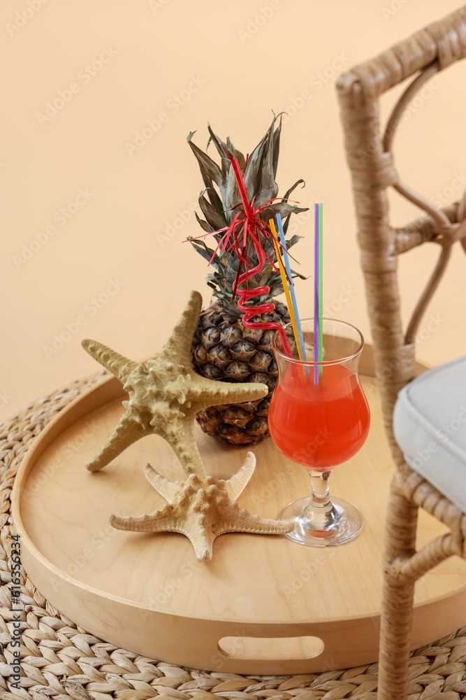 Wooden tray with cocktail, pineapple and starfishes on wicker ottoman against pale orange background