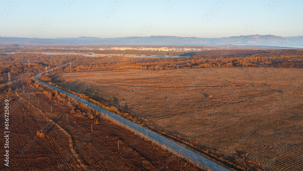 Autumn aerial photograph of the road among the fields. Tractors mow hay in the fields. A small town 