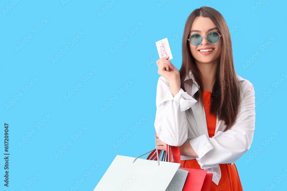 Young woman with gift card and shopping bags on blue background