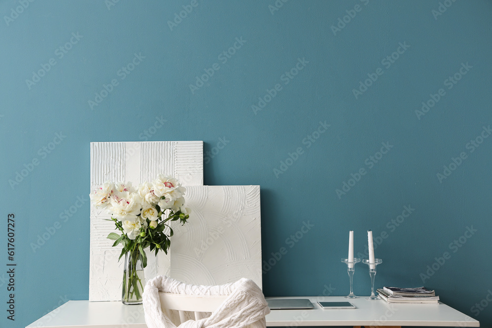 Vase of white peonies and paintings on table near blue wall