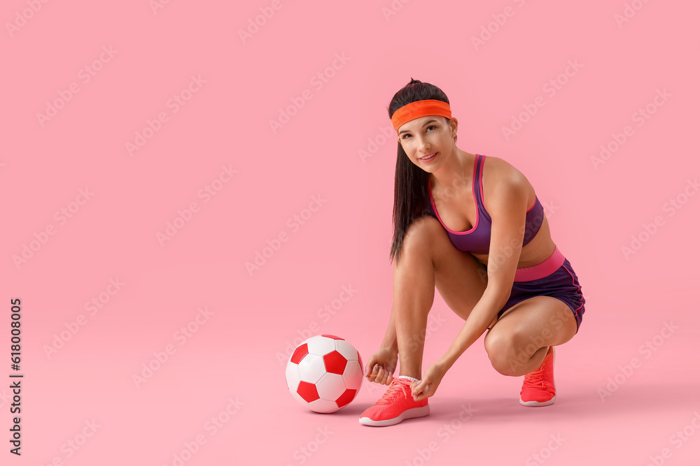 Sporty young woman with soccer ball tying shoelaces on pink background
