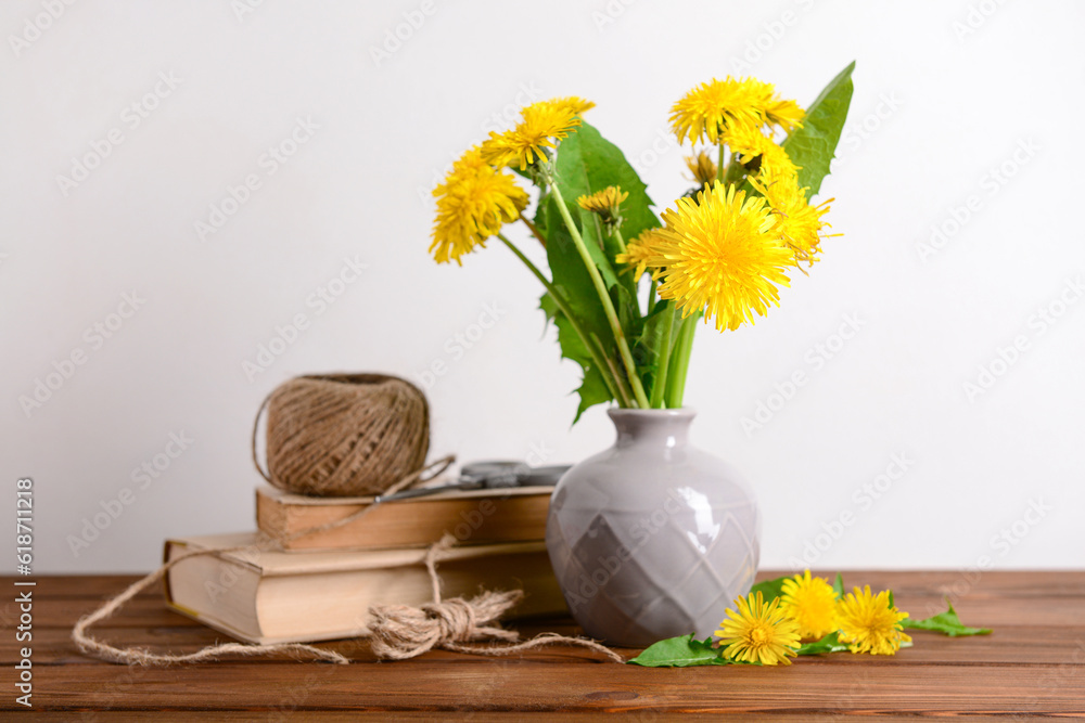 Vase with yellow dandelion flowers, rope, scissors and books on wooden table near light wall