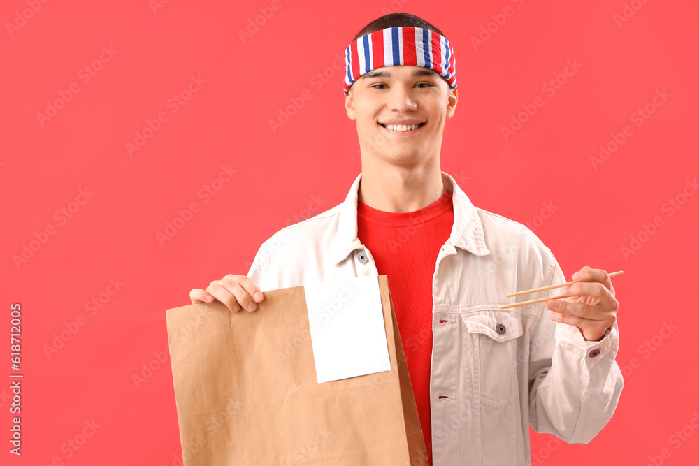 Young man holding paper bag with Asian food and chopsticks on red background