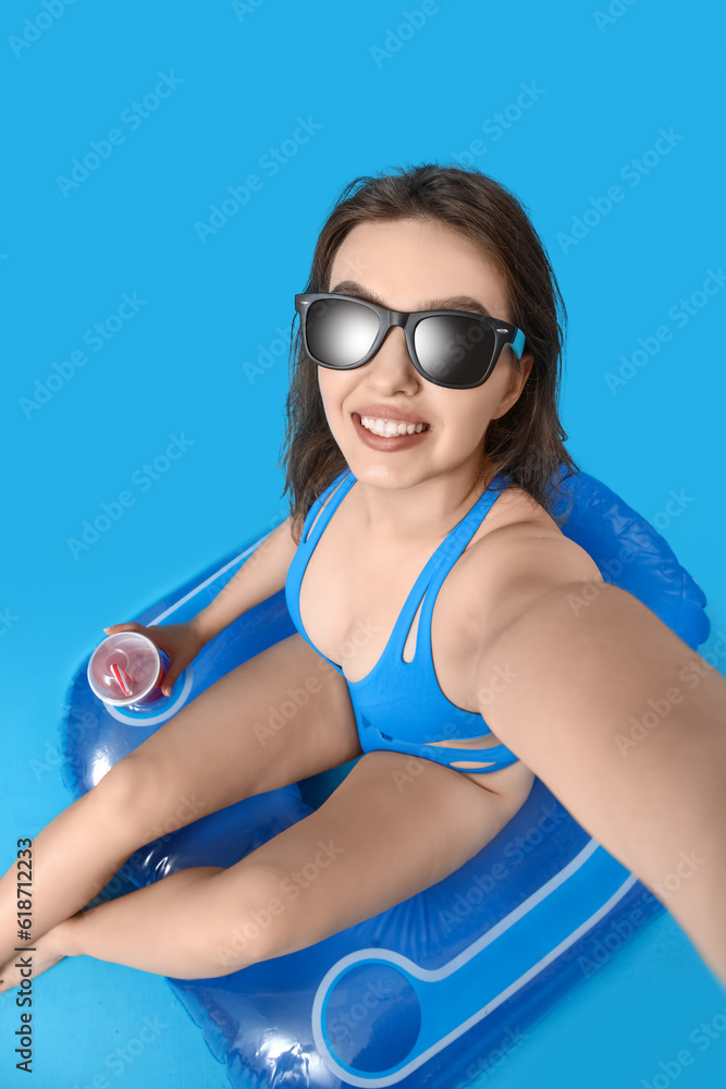 Young woman in sunglasses with swim ring taking selfie on blue background