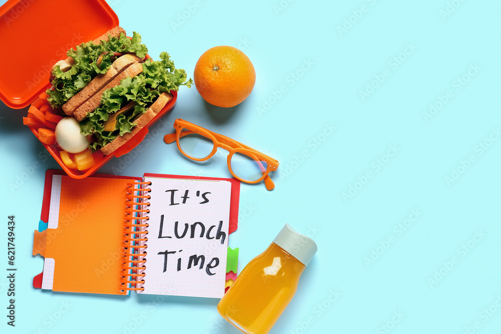 Notebook with text ITS LUNCH TIME, eyeglasses and tasty food in lunchbox on blue background