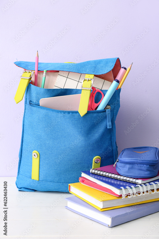 Blue school backpack with books, pencil case and markers on white wooden table near lilac wall