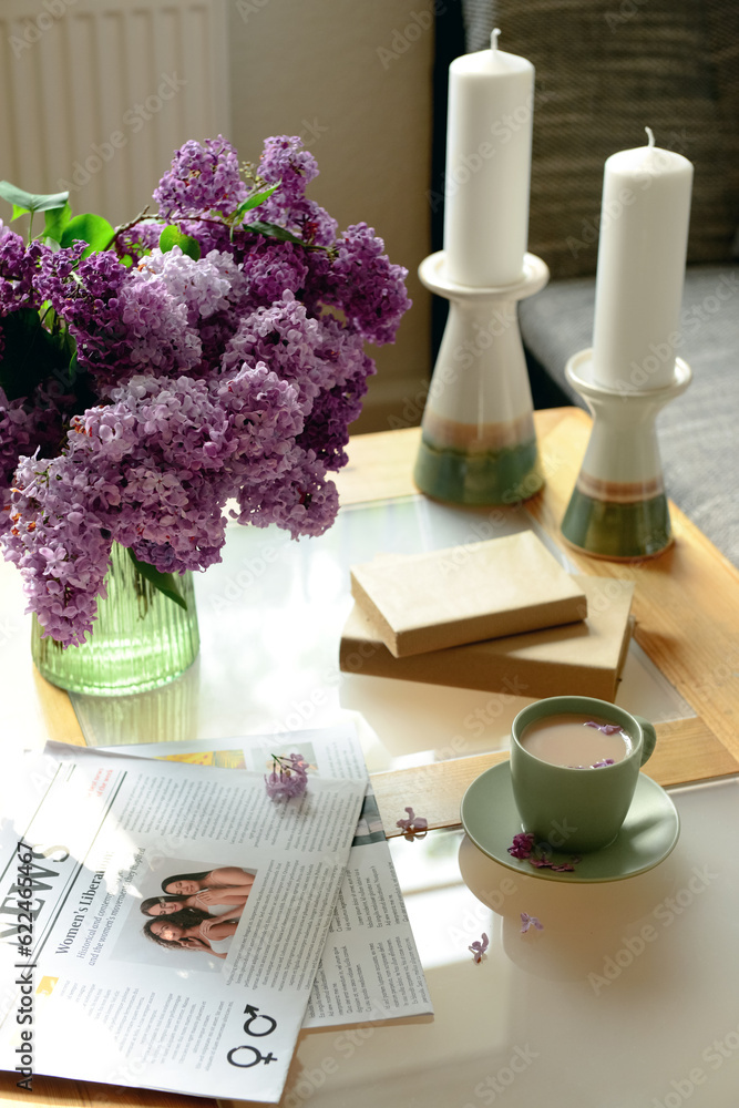 Vase with beautiful lilac flowers, candles, cup of coffee and newspaper on table in living room, clo