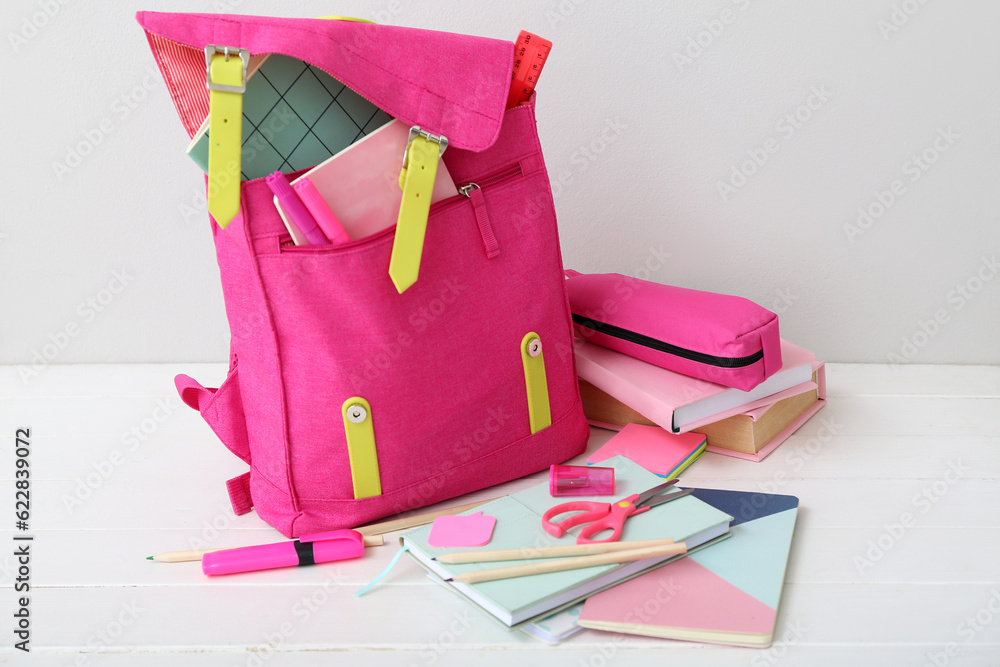 Pink school backpack with books, pencil case and stationery on white wooden table near wall