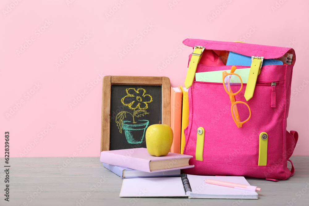 Pink school backpack with books, chalk drawing flower and apple on grey wooden table near color wall