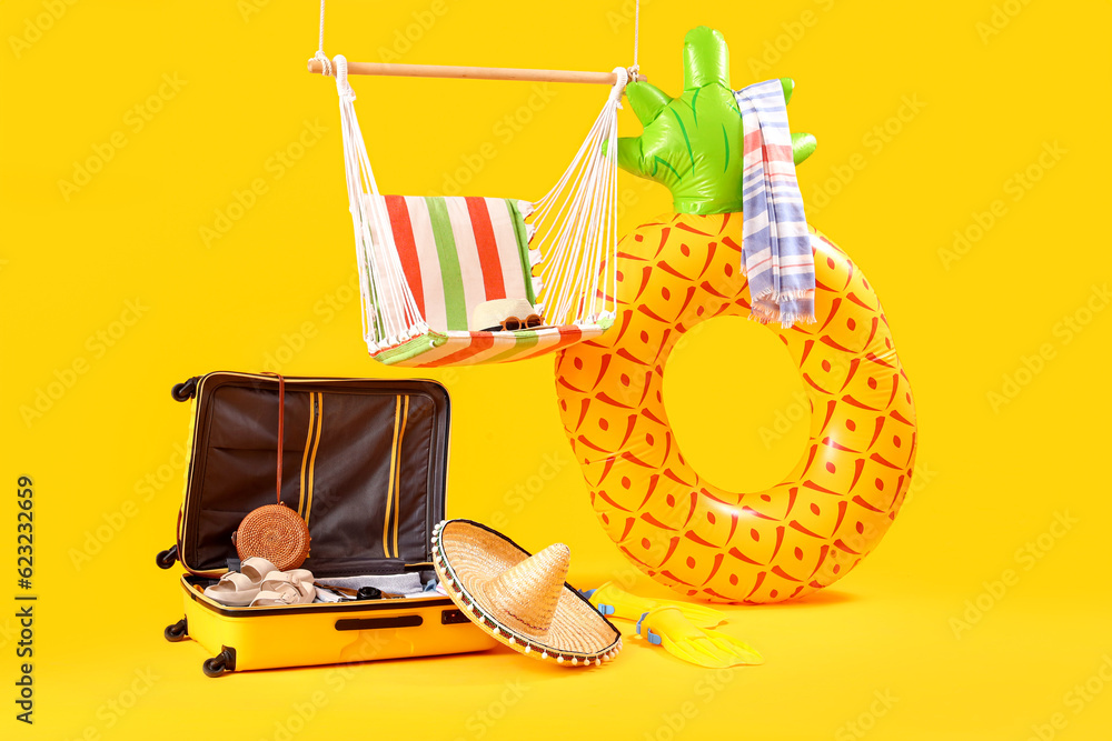 Hanging hammock with suitcase and beach accessories on yellow background. Travel concept
