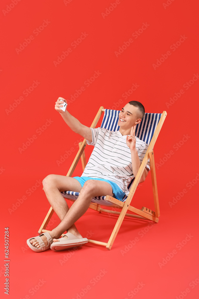 Happy young man sitting on deckchair and taking selfie against red background