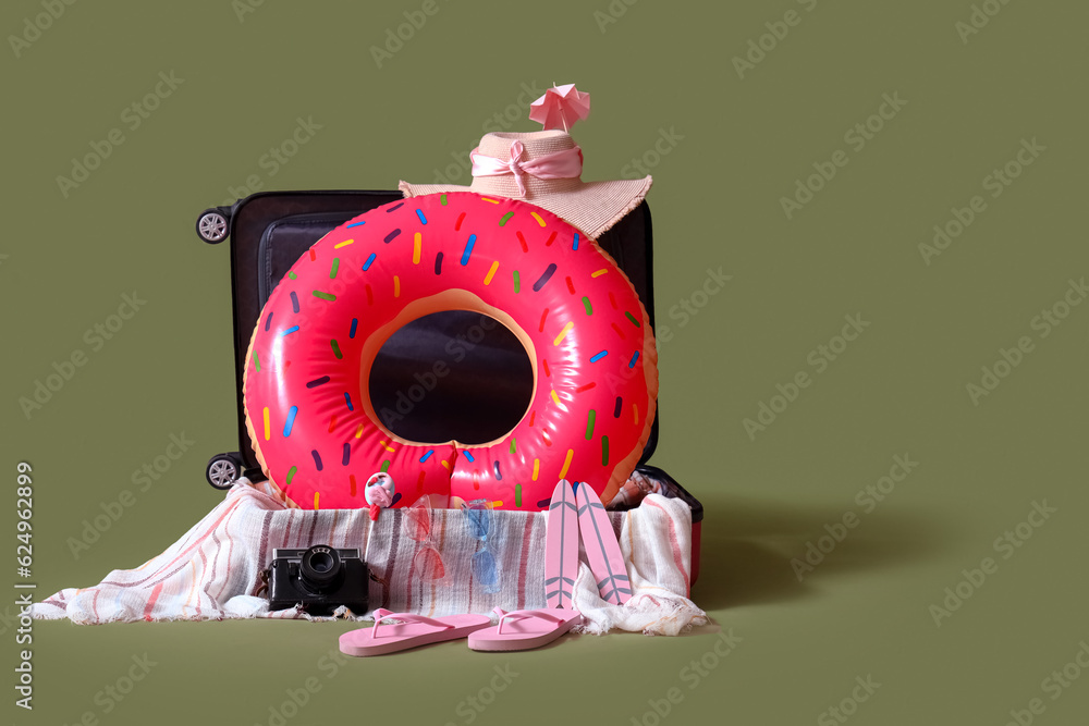Suitcase with inflatable ring in shape of donut and beach accessories on green background. Travel co
