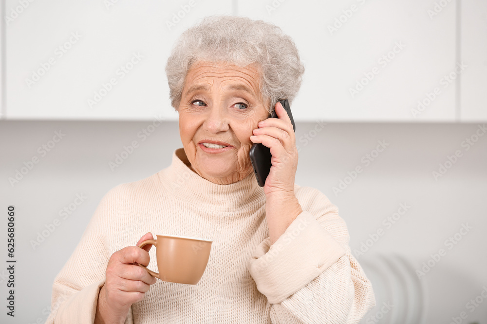 Senior woman with cup of tea talking by mobile phone in kitchen