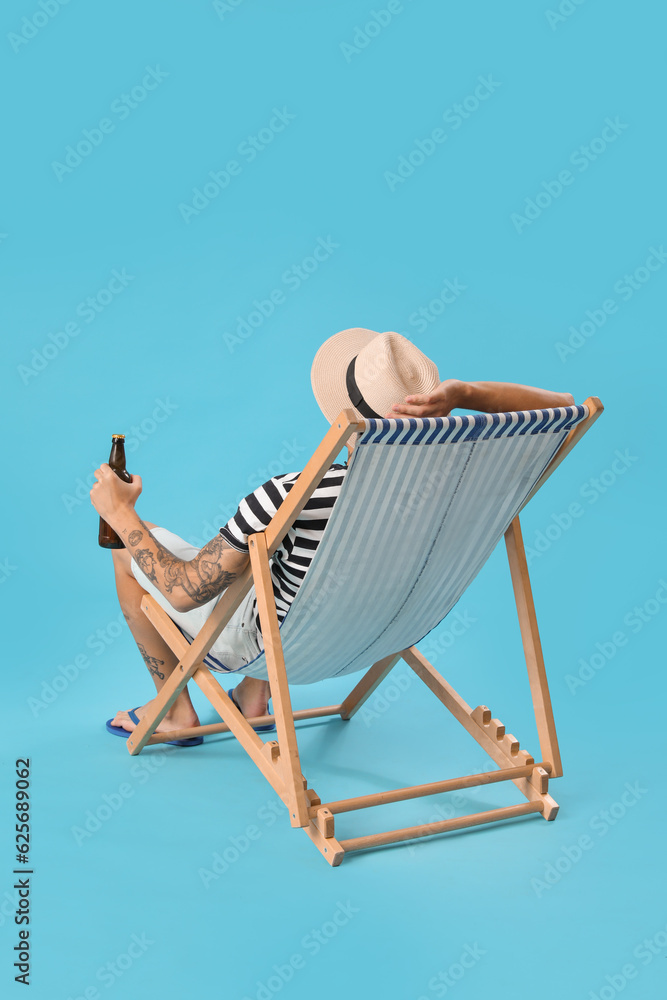 Young man with beer sitting in deck chair on blue background, back view