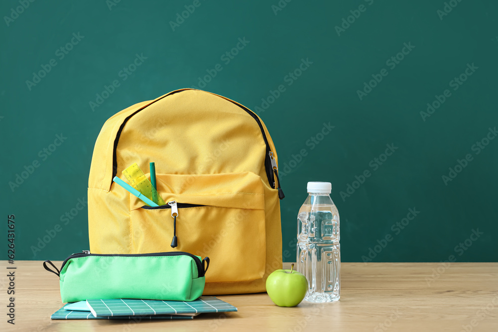 Yellow school backpack with stationery and bottle of water on wooden table near green chalkboard