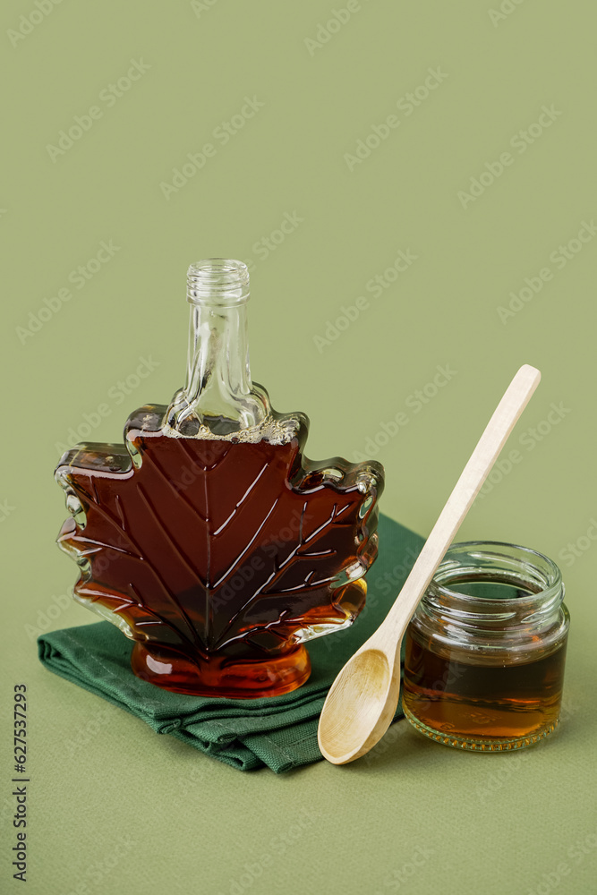 Bottle and jar with spoon of tasty maple syrup on green background