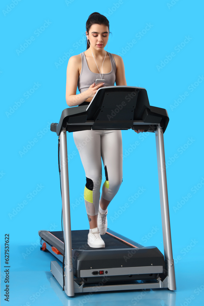 Sporty young woman with earphones and mobile phone training on treadmill against blue background