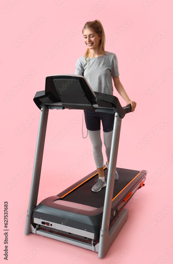 Sporty young woman training on treadmill against pink background