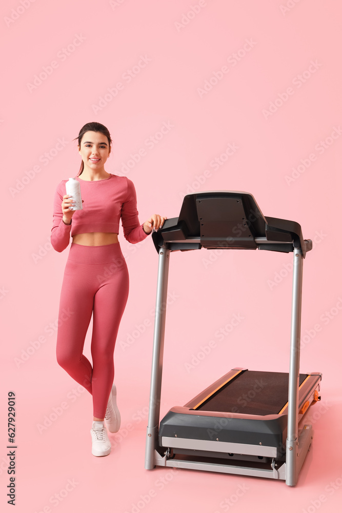 Sporty young woman with bottle of water and treadmill on pink background