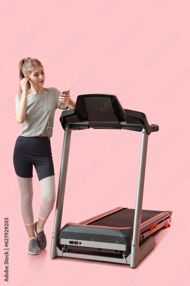 Sporty young woman listening to music near treadmill on pink background