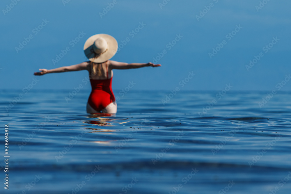 Beautiful woman in red swimsuit and hat standing in the sea, front focus on waves, blurred body
