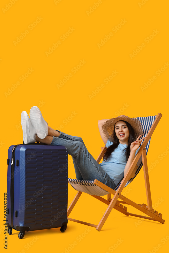 Young woman with suitcase in deck chair showing victory gesture on yellow background