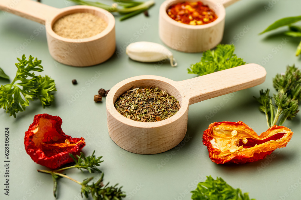 Wooden scoops with spices, dried tomatoes and herbs on color background, closeup