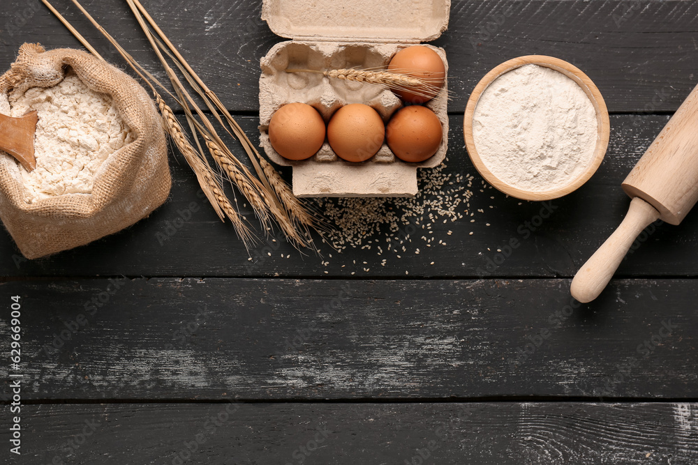 Composition with flour, wheat ears, eggs and rolling pin on black wooden table