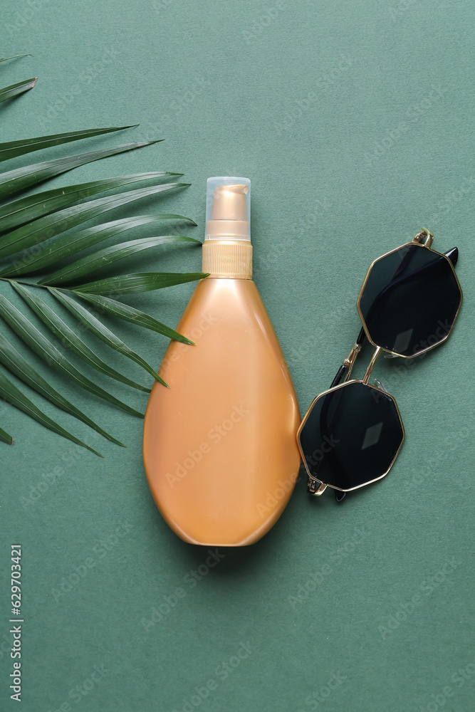 Bottle of sunscreen cream, sunglasses and palm leaf on green background