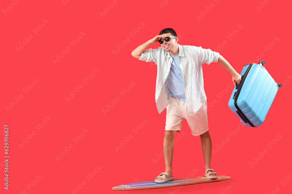 Male tourist with suitcase standing on surfboard against red background