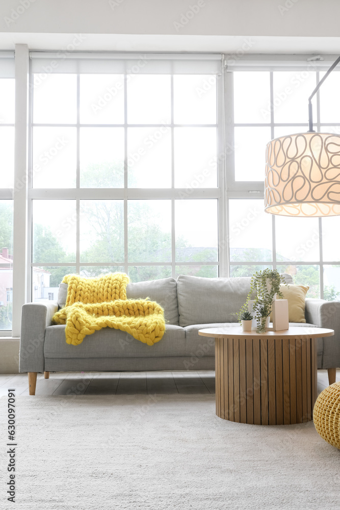 Interior of light living room with cozy grey sofa, coffee table and glowing lamp near big window