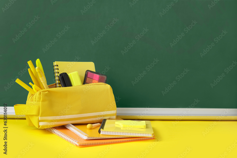Pencil case with different school stationery on yellow table near blackboard