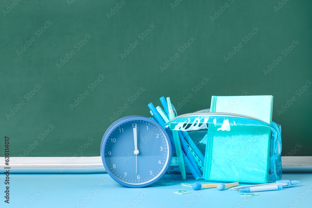 Pencil case with different school stationery and alarm clock on blue table near blackboard